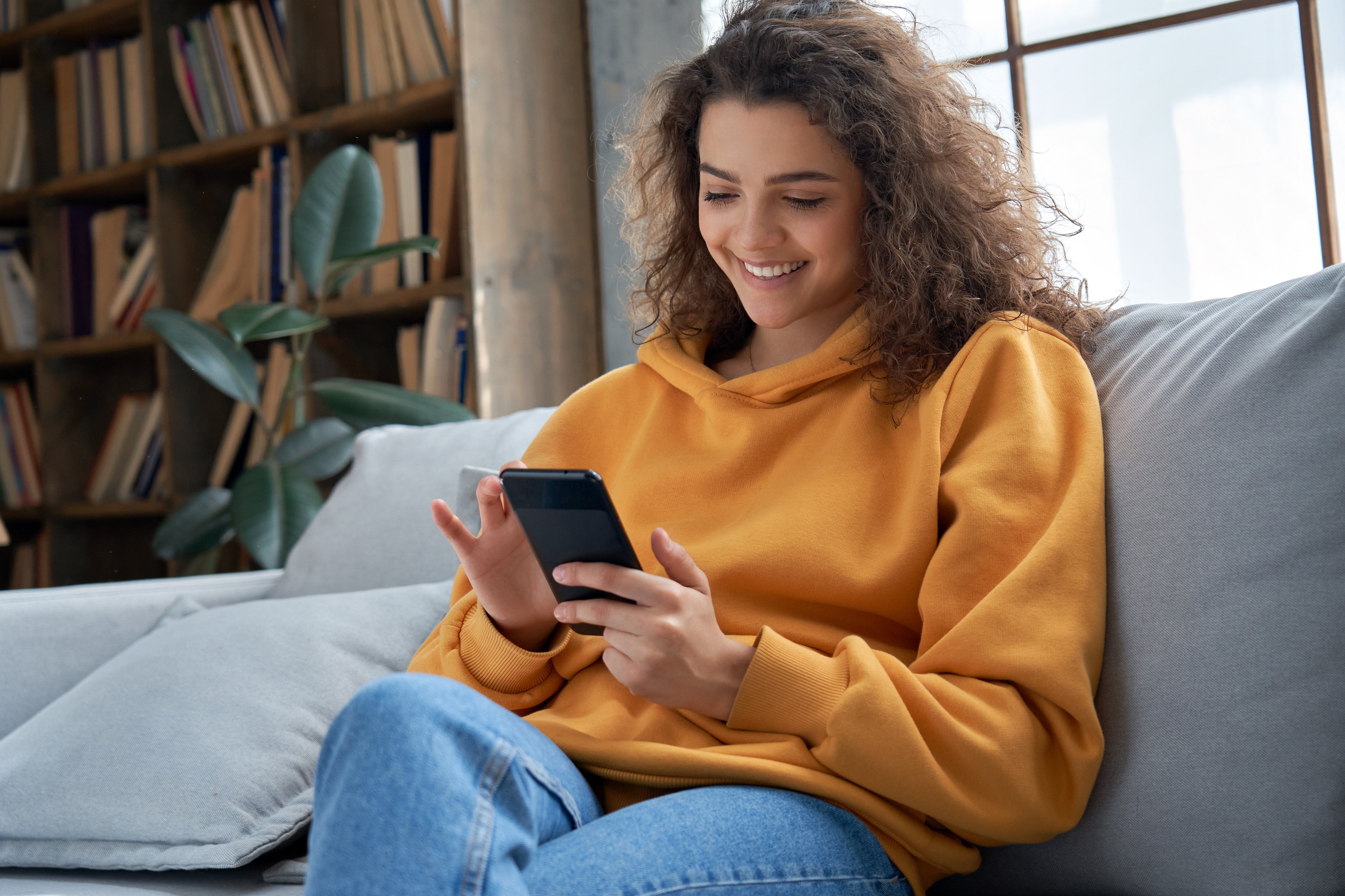 Millennial girl sitting on the couch holding and smiling at her phone.