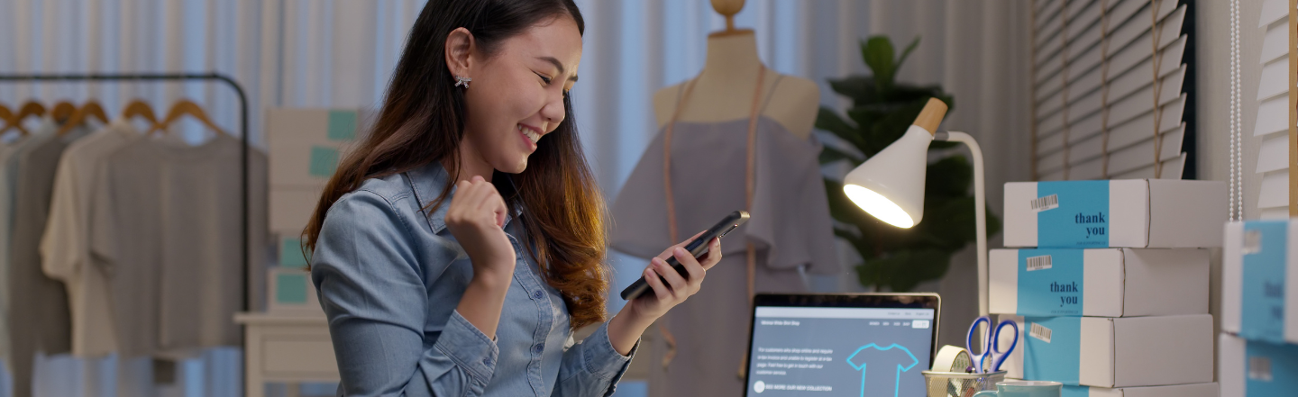 A girl smiling holding her phone, a laptop and boxes on her desk.