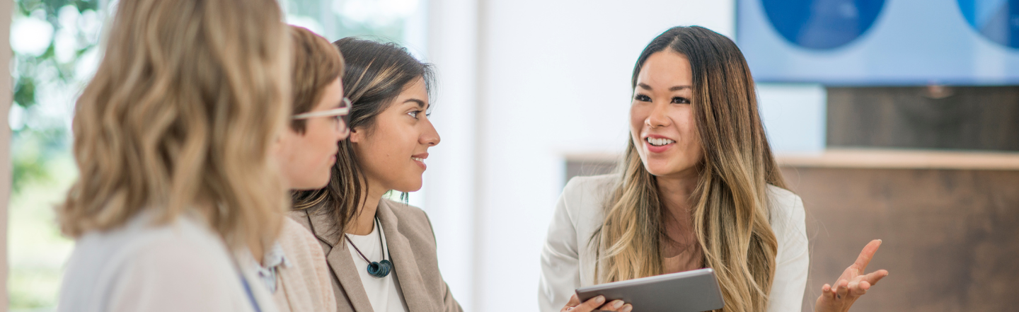 A confident and cheerful woman leading a discussion with three other women in a modern, well-lit office space. She is holding a tablet and gesturing while speaking, conveying collaboration and connection.