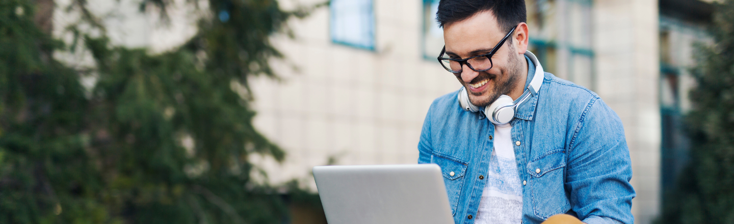 A man wearing glasses and a denim shirt is smiling while working on a laptop outdoors. He has headphones around his neck, and the background features trees and a blurred building, creating a calm and relaxed atmosphere.
